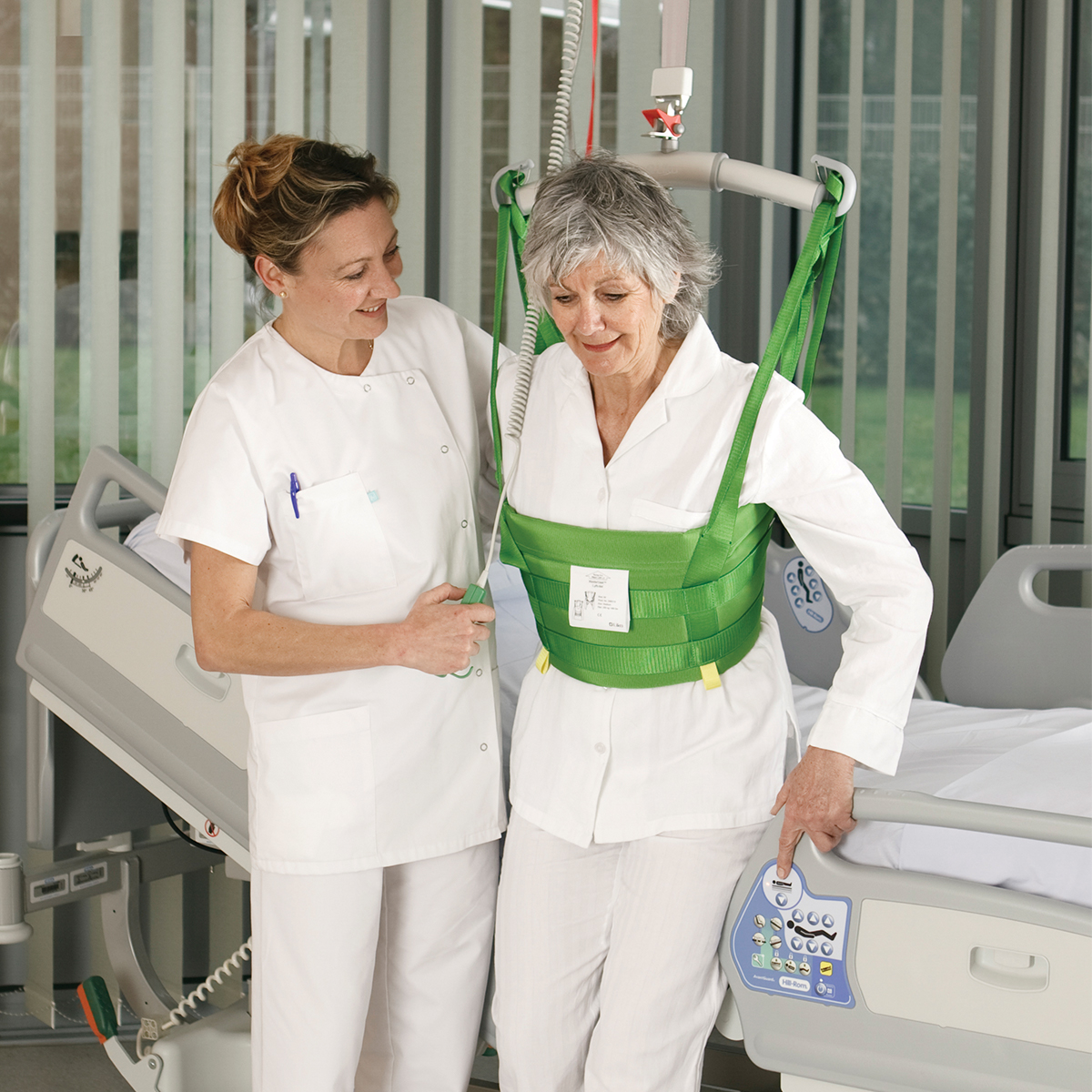 Un profesional de la salud ayuda a un paciente a caminar en una habitación de hospital con una grúa de techo Hillrom y MasterVest